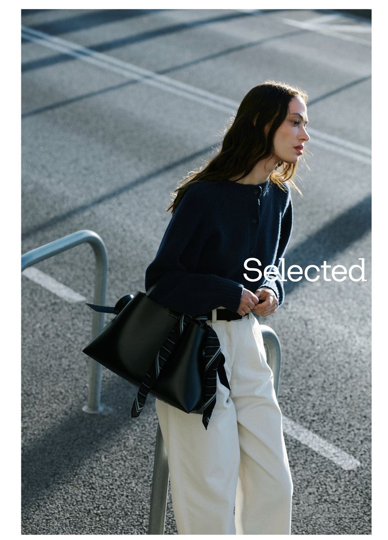 Model in navy sweater and white trousers leaning against metal rails with a black handbag