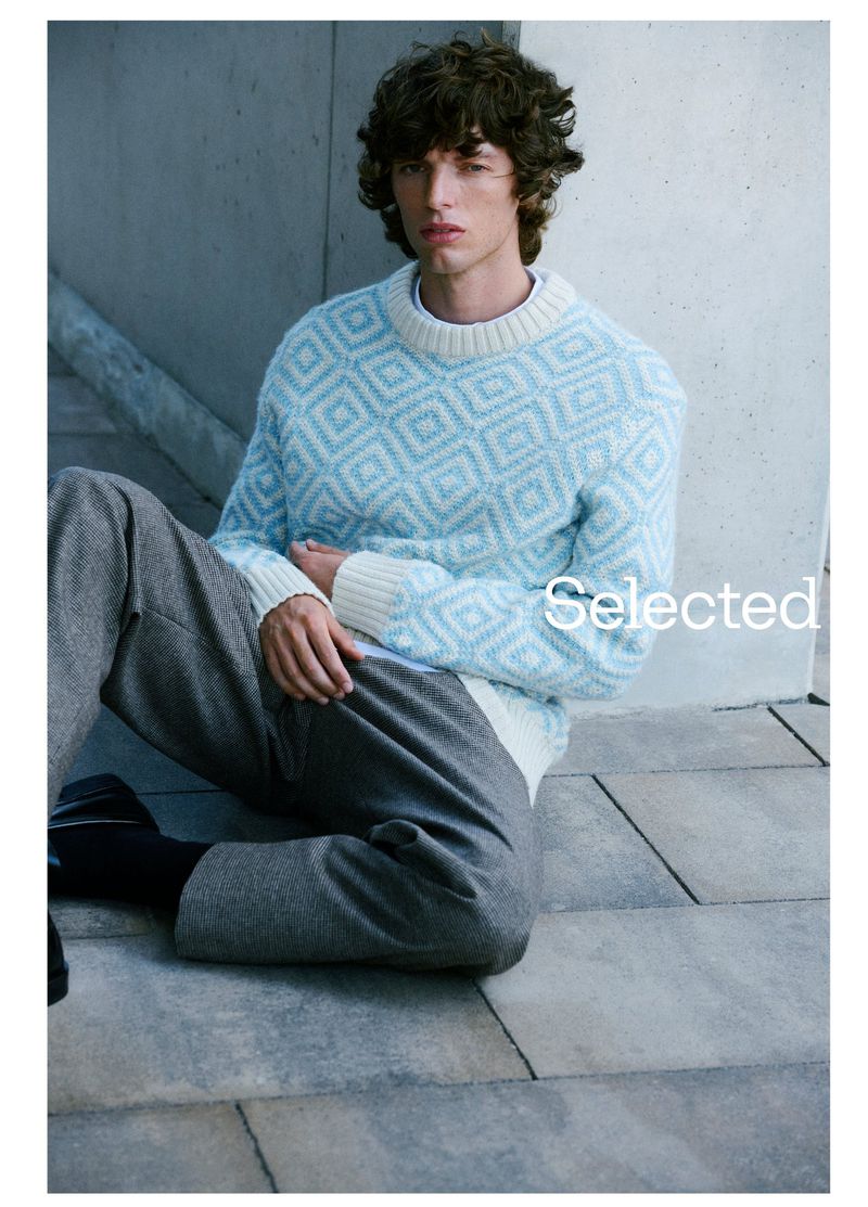 A young adult male model wearing a light blue geometric sweater and grey pants poses indoors against a grey concrete wall.