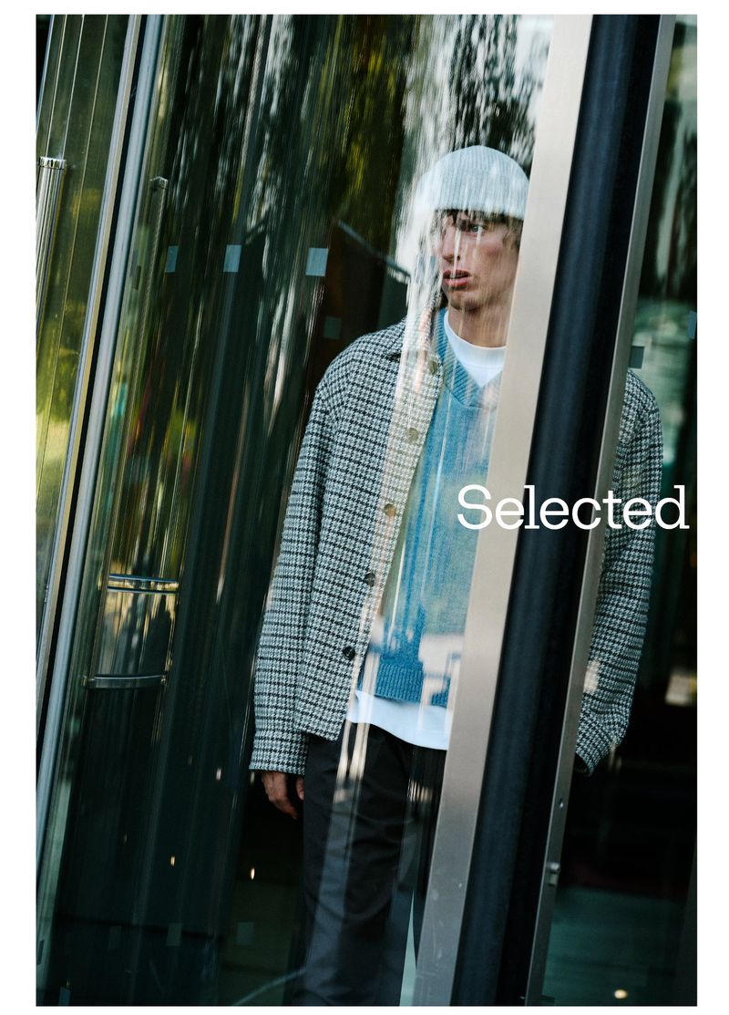 Male fashion model standing behind a store window, wearing a jacket, sweater, t-shirt and pants. Outdoor setting with sunlight.