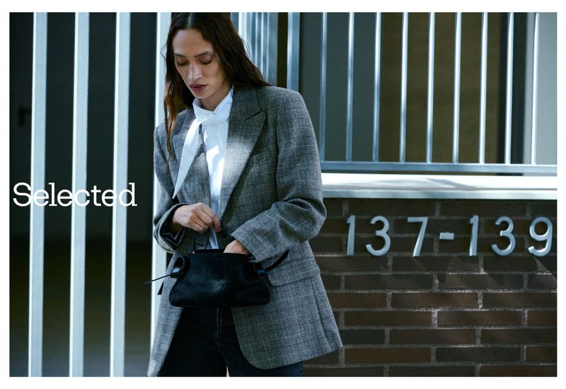 A woman in a grey blazer and white bow blouse holds a black purse, posing in front of a textured brick wall with silver numbers.