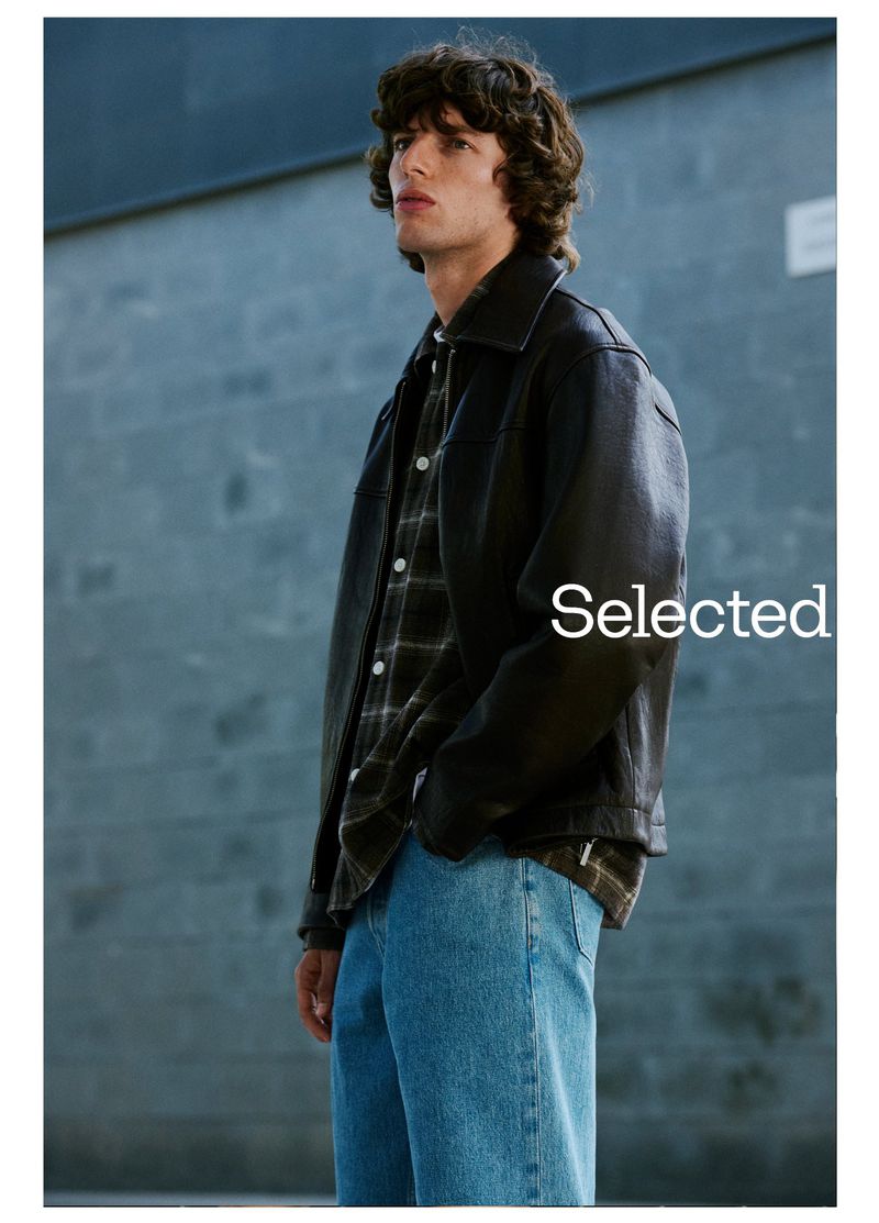 A young adult male poses in a dark jacket and jeans against a grey wall in a fashion-style full shot, with the brand 'Selected' overlaid.