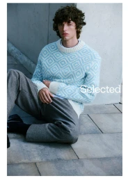 Male model seated on stone paving wearing a light blue patterned knit sweater and grey trousers for Selected campaign