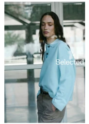 Model in a light blue polo sweatshirt and dark trousers standing before a large glass window for Selected FW Campaign.