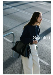 Model in navy sweater and white trousers leaning against metal rails with a black handbag