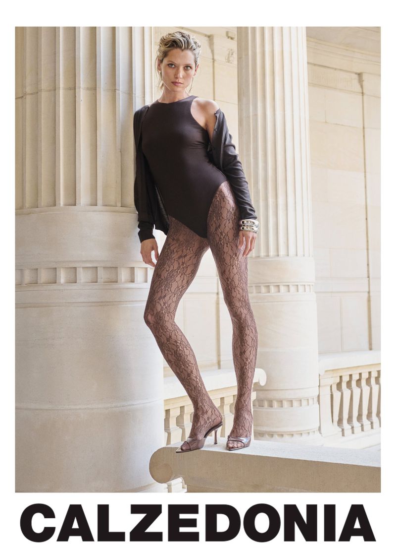 Indoor fashion shot of a woman wearing a brown bodysuit, lace tights, open jacket, and heels standing next to stone columns.