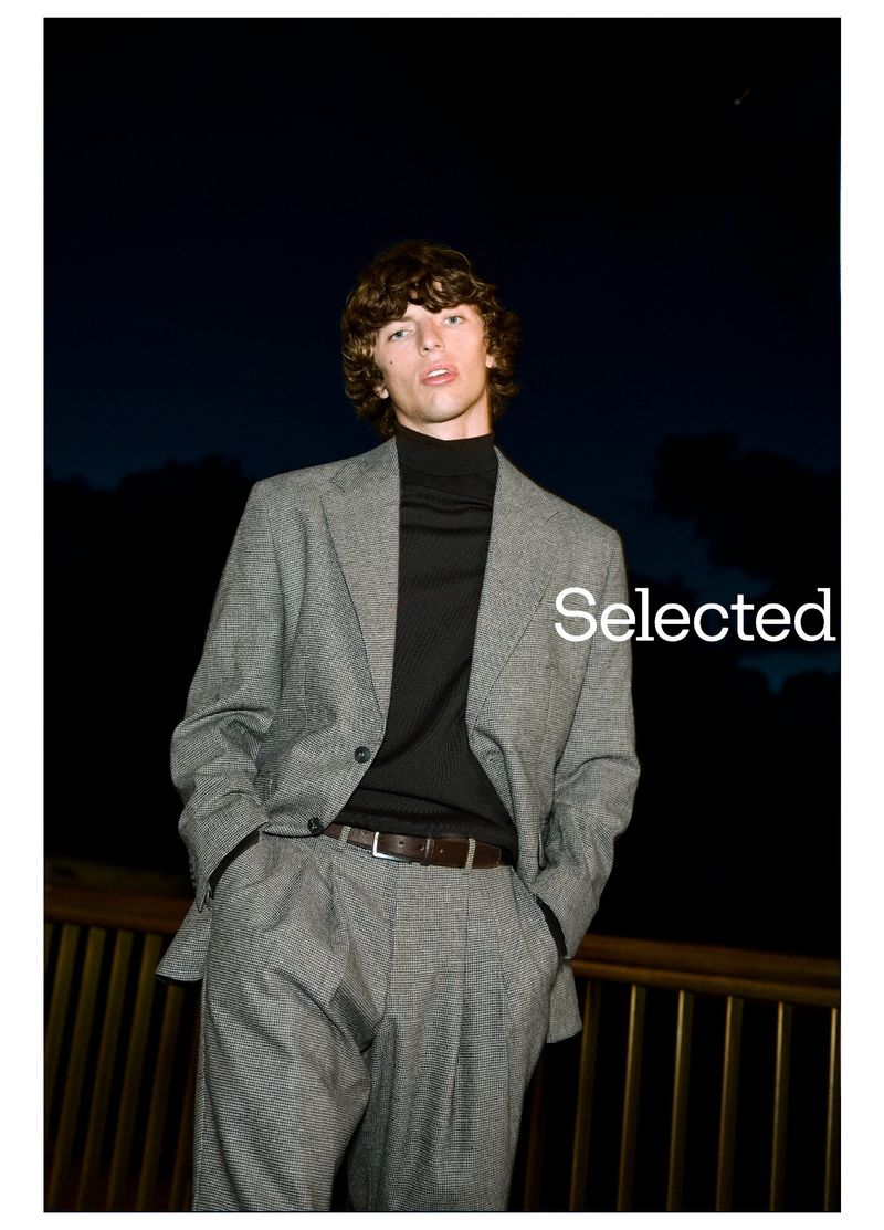 Male fashion model poses in a gray suit with a black turtleneck against a dark background, showcasing a blend of classic and contemporary style.