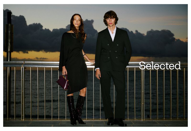 Two fashion models pose outdoors at twilight. The woman wears a dark dress and shiny boots. The man is in a dark suit.