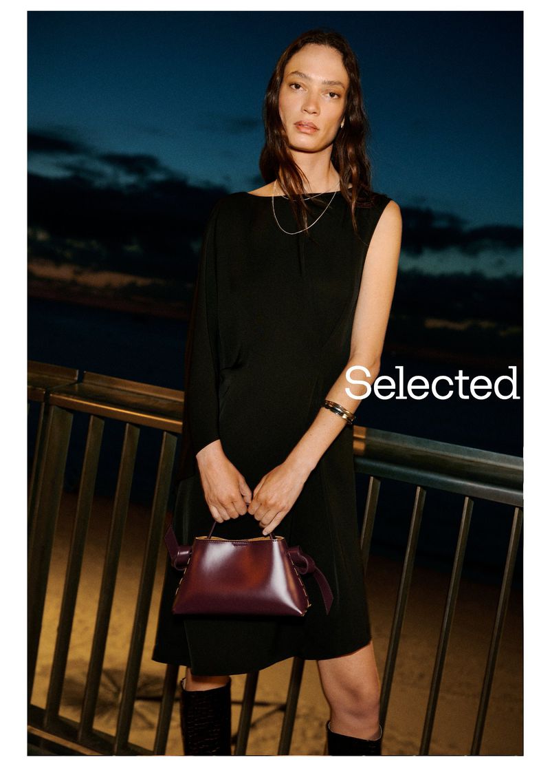 A brunette woman poses in a black dress and knee-high boots, holding a burgundy top-handle bag against a dark blue sky background.