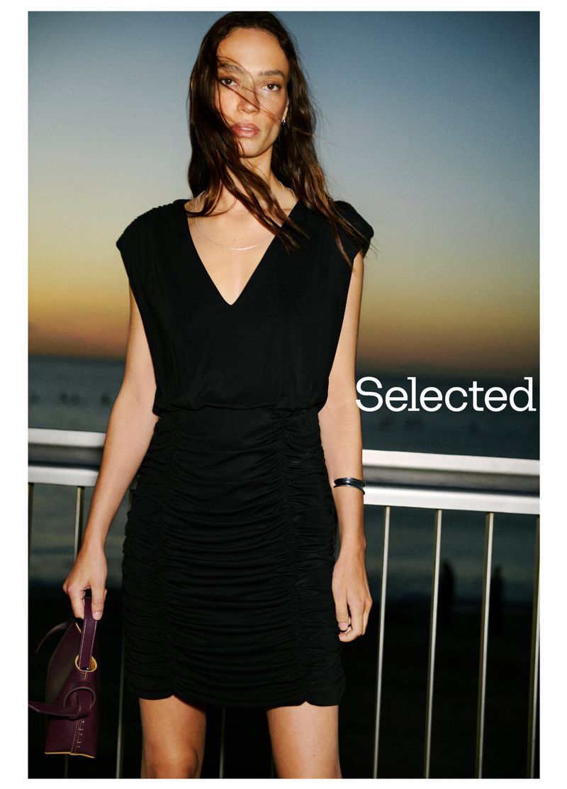 Woman wearing a short black dress and holding a handbag, posing against a dusky sky and blurred ocean backdrop.
