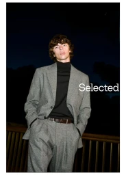 Man wearing a grey suit jacket and trousers with a black turtleneck standing outdoors at night.