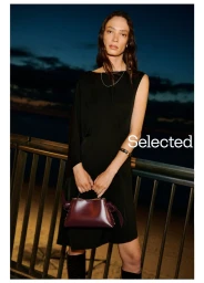 Model in a black dress holding a burgundy handbag standing by a railing at dusk for Selected Selected Holiday Campaign