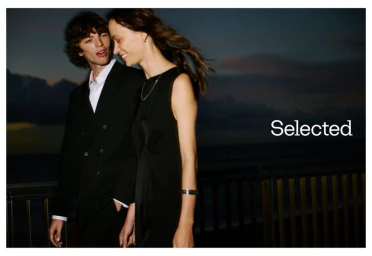 Two models outdoors at dusk, a man in a black blazer and a woman in a black dress, with the brand name Selected visible.