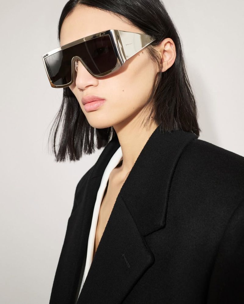 Portrait of a woman wearing oversized gold sunglasses and a black wool coat against a neutral backdrop in a fashion editorial style.