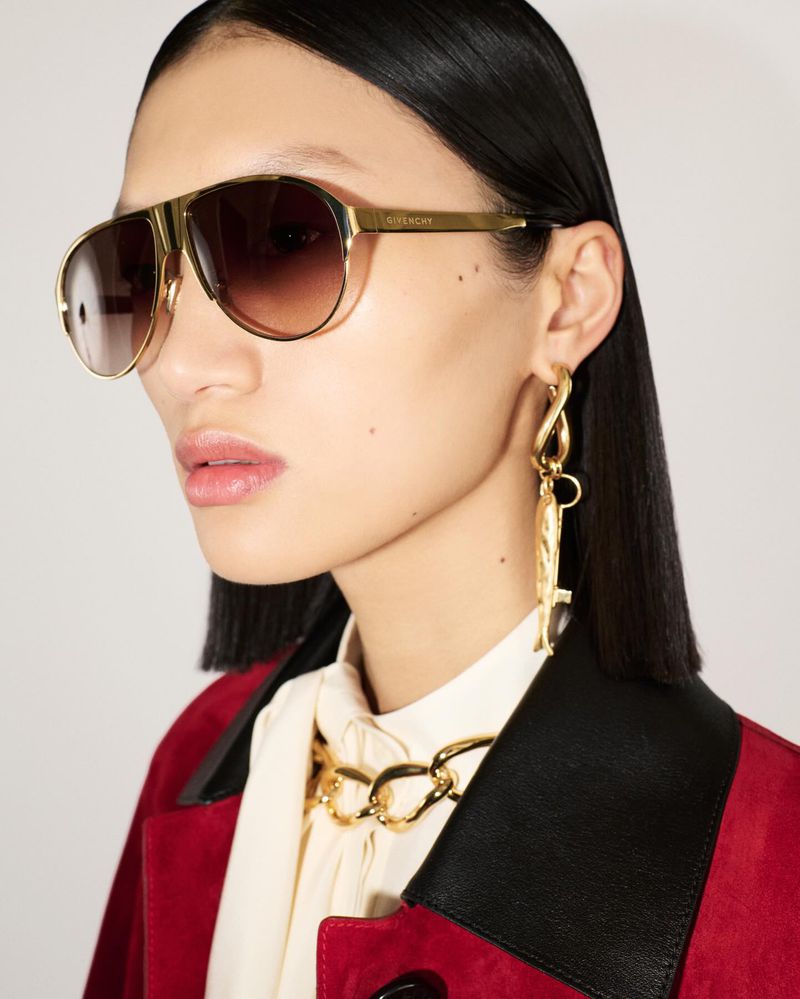 Model wearing gold aviator sunglasses, sculptural earrings, and a red coat against a white background for Givenchy.