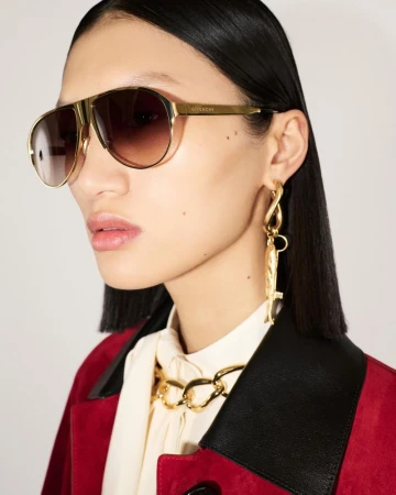 Model wearing gold aviator sunglasses, sculptural earrings, and a red coat against a white background for Givenchy.