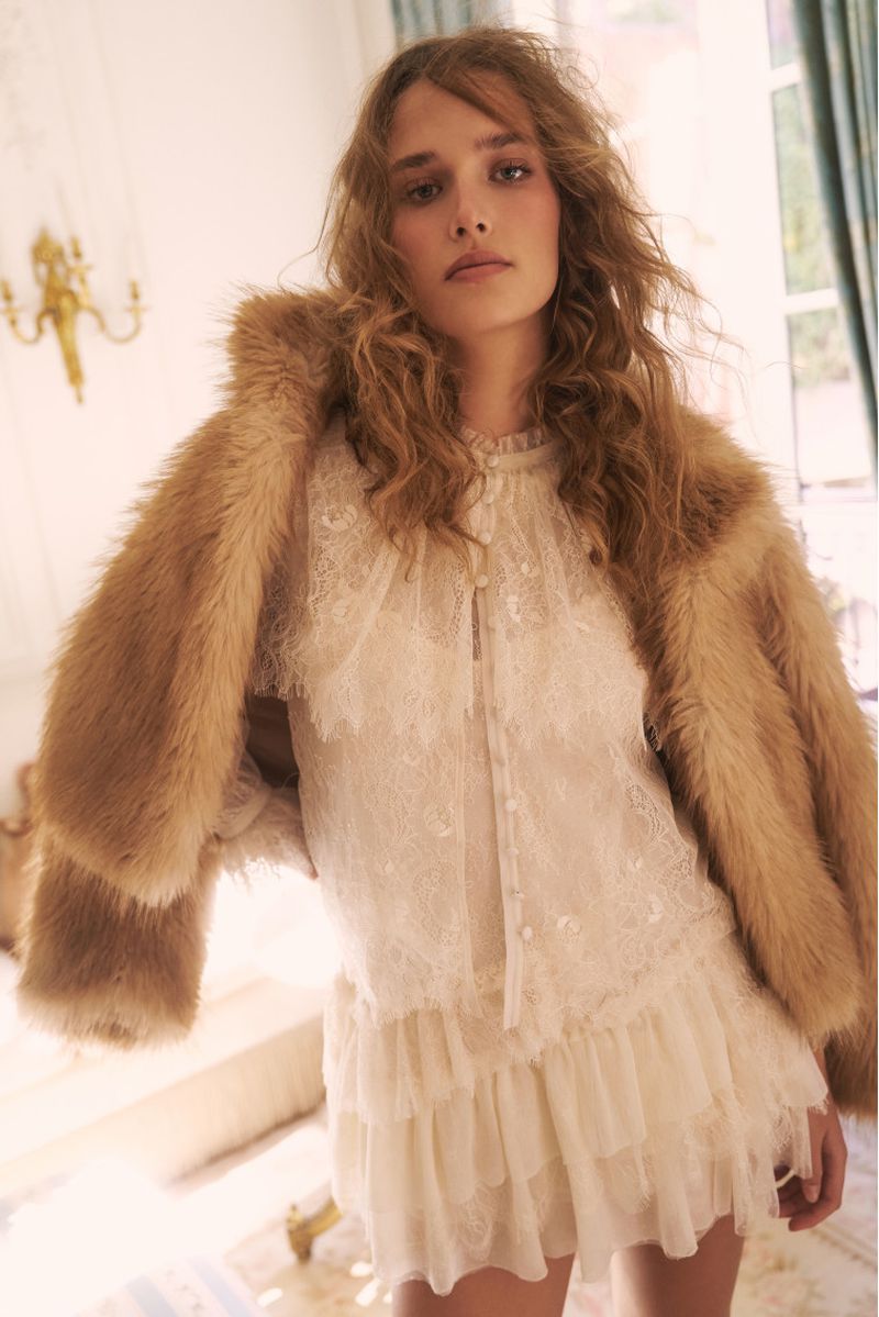 A woman with curly hair stands indoors wearing a short white lace dress with a fur jacket, illuminated by natural light and a golden wall sconce.