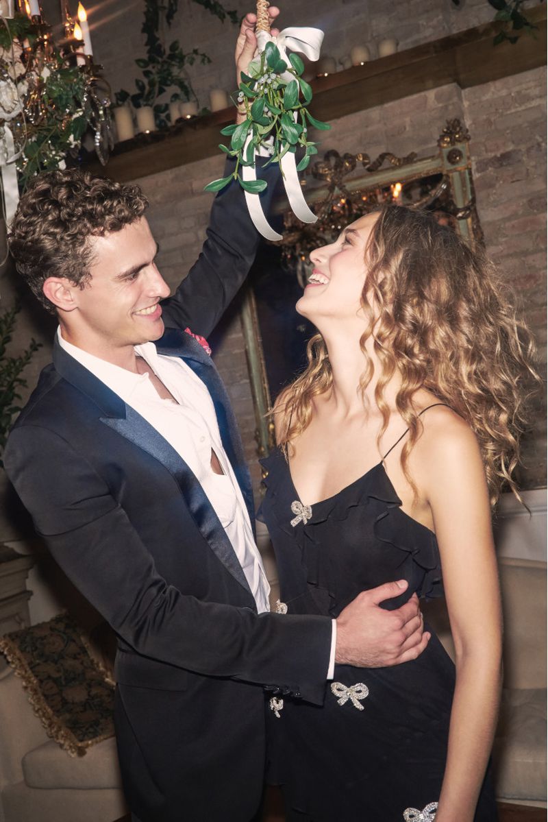 A smiling couple in formalwear stands indoors under mistletoe; the man wears a tuxedo, and the woman wears a black dress with ribbon details.
