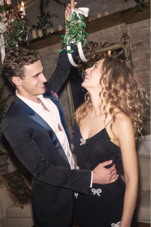 A smiling couple in formalwear stands indoors under mistletoe; the man wears a tuxedo, and the woman wears a black dress with ribbon details.