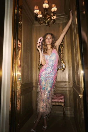 Woman in shimmering gown poses in paneled interior with chandelier, holding masquerade mask and reaching arm up in elegant fashion shot.