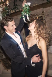 A man in a dark suit holding mistletoe above a smiling woman in a black dress for Loveshackfancy Holiday '24 campaign