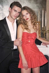 Man in a black tuxedo and woman in a red strapless bow dress posing together in an elegant interior with a fireplace and lit candles.