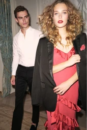 A woman in a red ruffled dress with a black blazer draped over her shoulders, accompanied by a man in a white shirt and dark trousers.