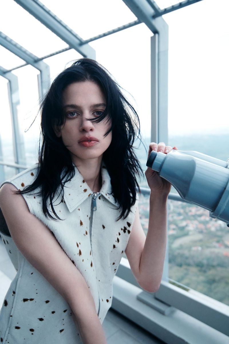 Young woman with dark hair and a vest-style shirt looks out from a skyscraper viewing platform, using binoculars to observe the cityscape.