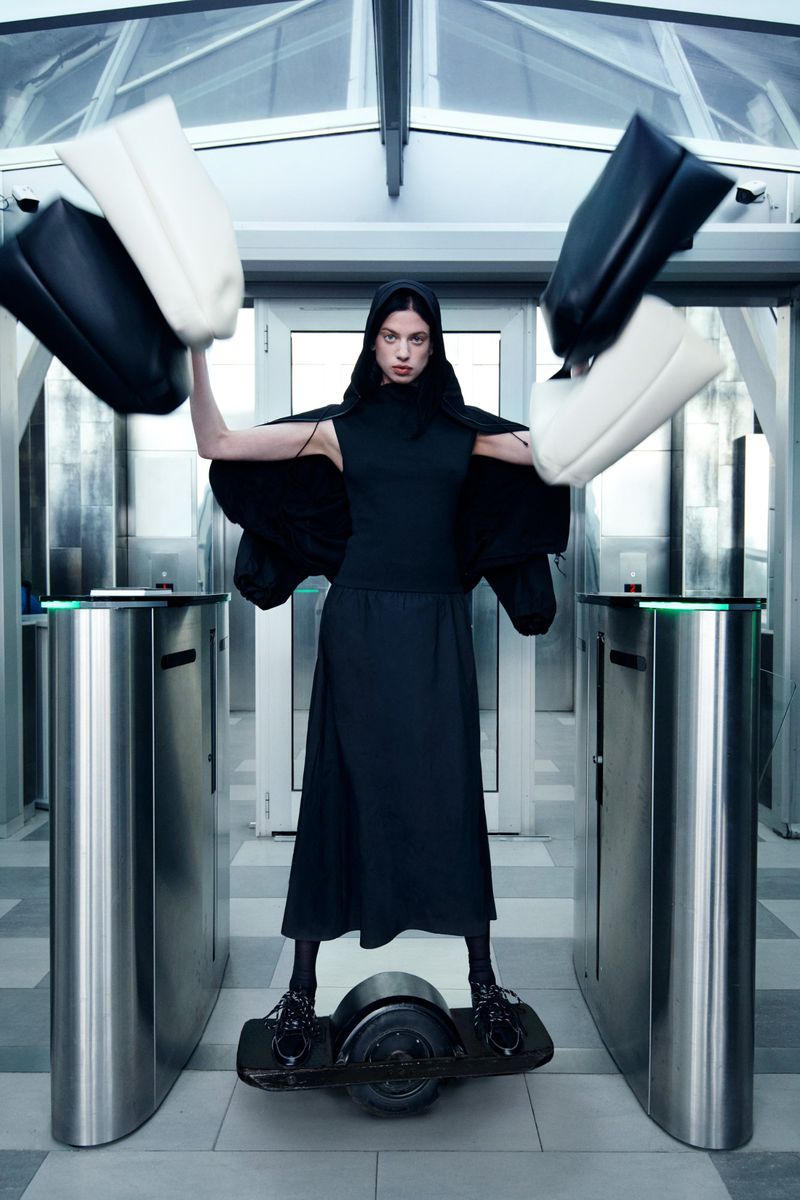 Fashionable woman in black outfit, wearing black platform shoes and riding an electric skateboard, holding two large handbags.