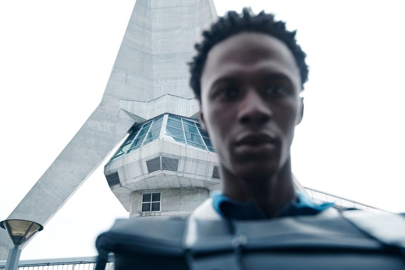 Portrait of a young man with short black hair in a dark top, set against a concrete tower with glass windows under a white sky.