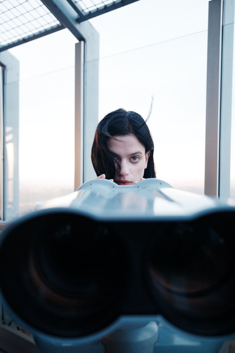 Woman poses behind coin-operated binoculars, gazing directly at the camera, with city structures visible through a window.
