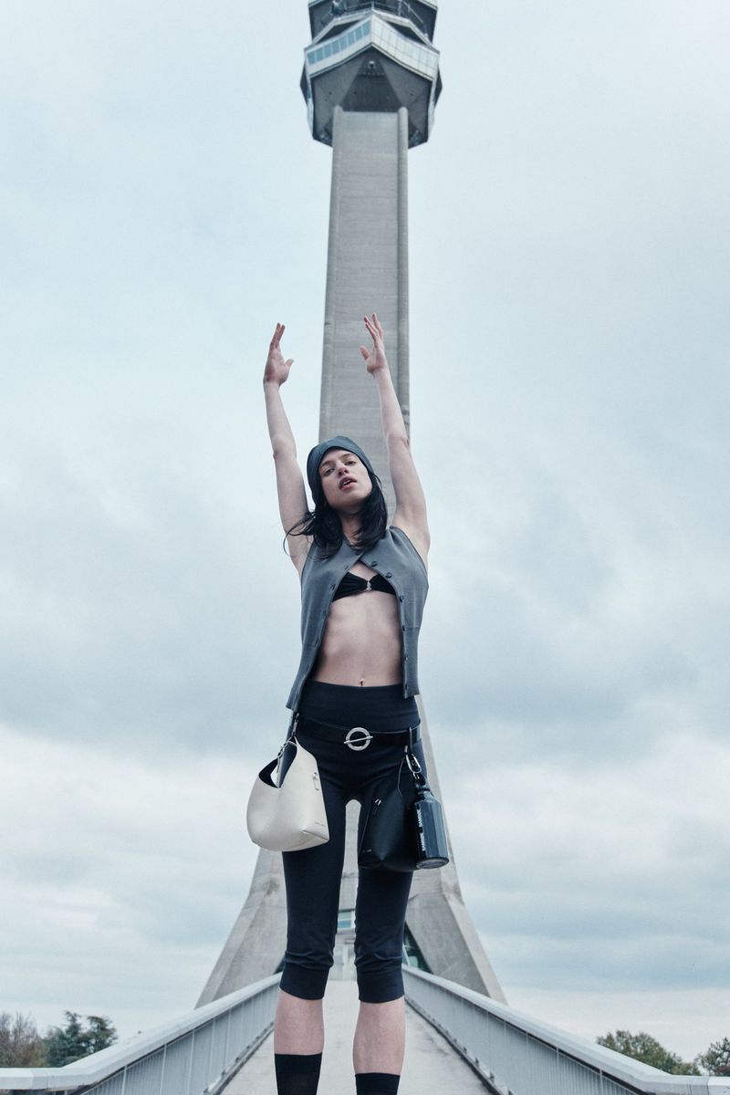Woman poses on a bridge with two bags, arms raised, against a backdrop of a tall tower and cloudy sky in an outdoor setting.