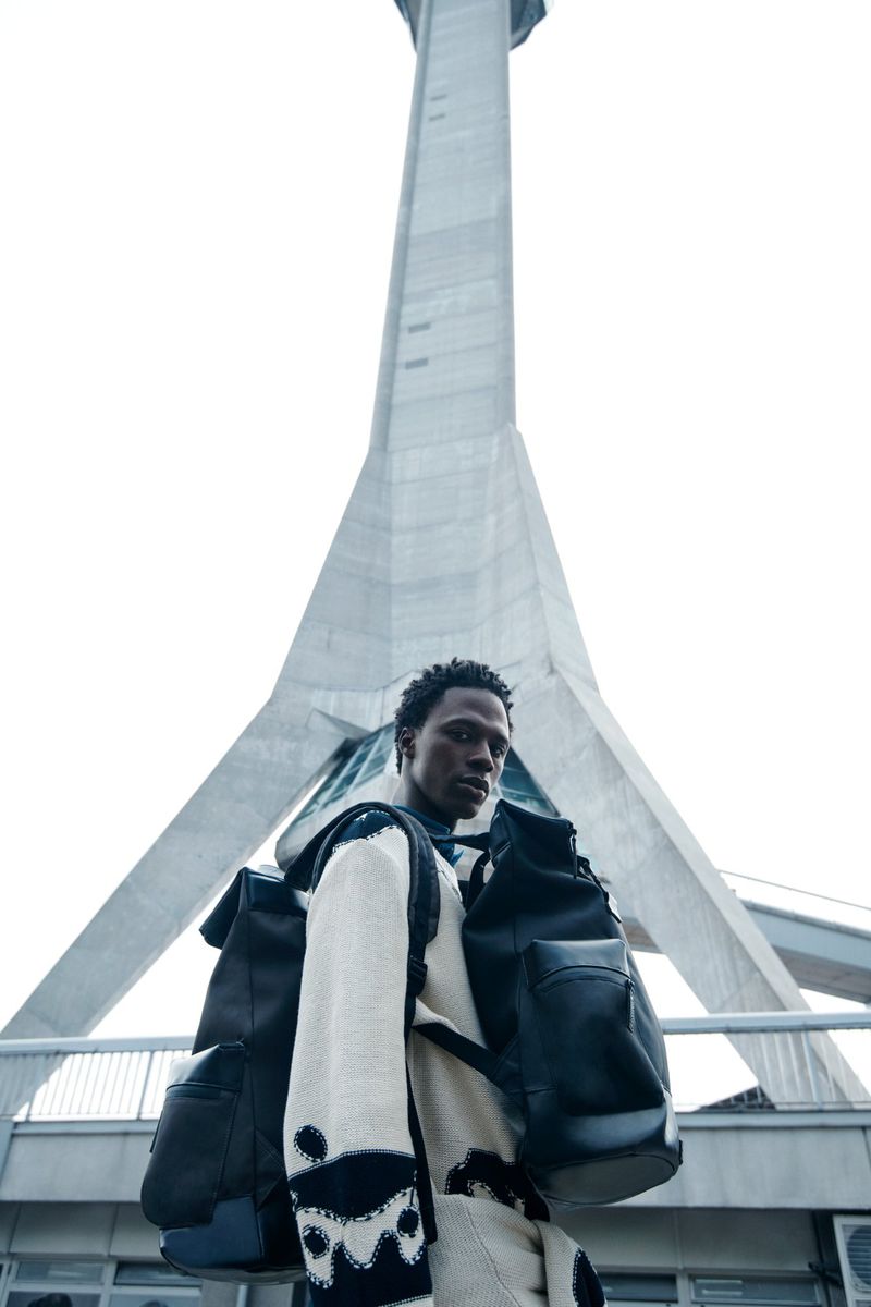 An adult male model in casual fashion stands in front of a tall, modern tower on an overcast day for an outdoor editorial fashion shot.