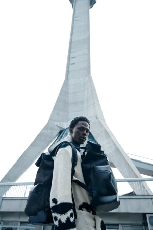 An adult male model in casual fashion stands in front of a tall, modern tower on an overcast day for an outdoor editorial fashion shot.