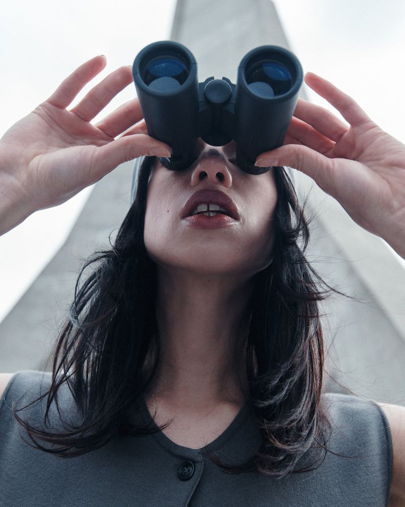 Low angle of a woman looking through binoculars, wearing a grey outfit against a bright, grey sky and a geometric building.