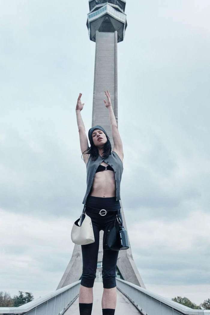 Model with arms raised stands on a bridge before a tall concrete tower for Samsøe Samsøe spring summer 2026 campaign.