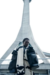 Model standing outdoors wearing a patterned sweater and carrying backpacks with a tall concrete tower in background
