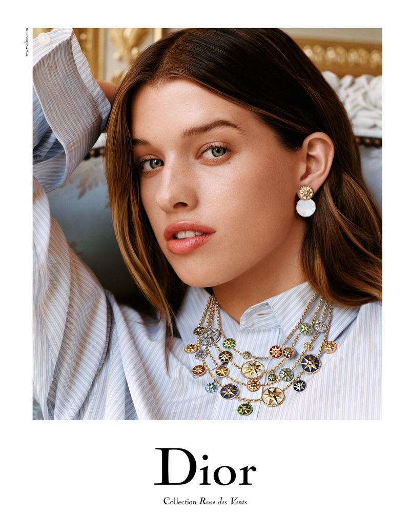 Close-up of an adult female model wearing a striped shirt, a pearl earring, and a layered gold necklace for a Dior jewelry advertisement.