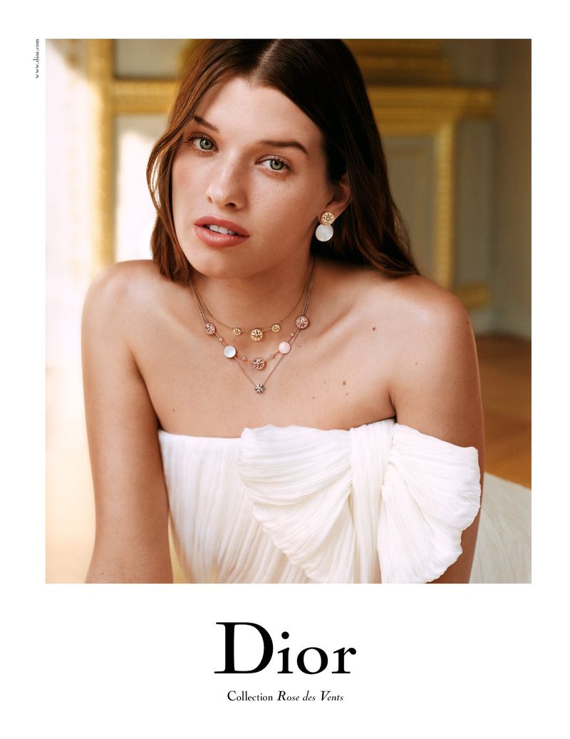 Model wearing Dior jewelry: a necklace and earrings, in a close-up shot. The model is fair-skinned and has light brown hair in soft waves.