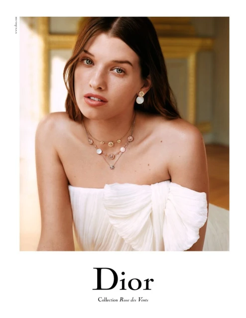 Model wearing Dior jewelry: a necklace and earrings, in a close-up shot. The model is fair-skinned and has light brown hair in soft waves.