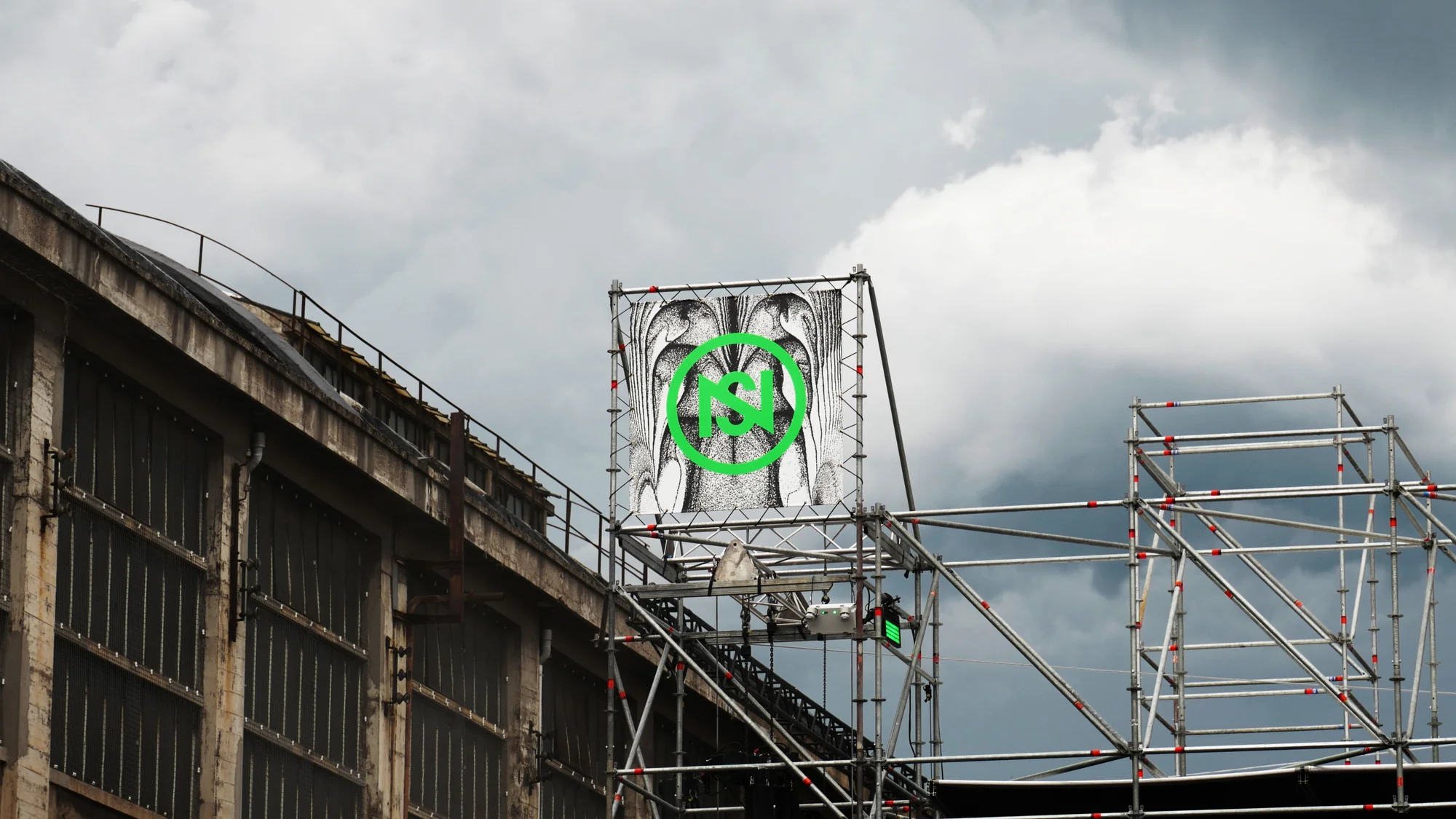 Artwork displayed on scaffolding next to an industrial building under a cloudy sky, showcasing urban art and structural elements.
