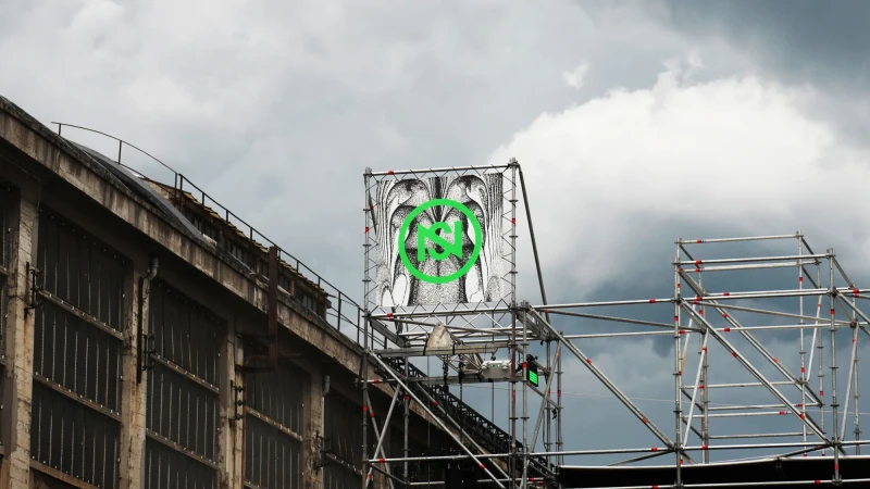 Artwork displayed on scaffolding next to an industrial building under a cloudy sky, showcasing urban art and structural elements.