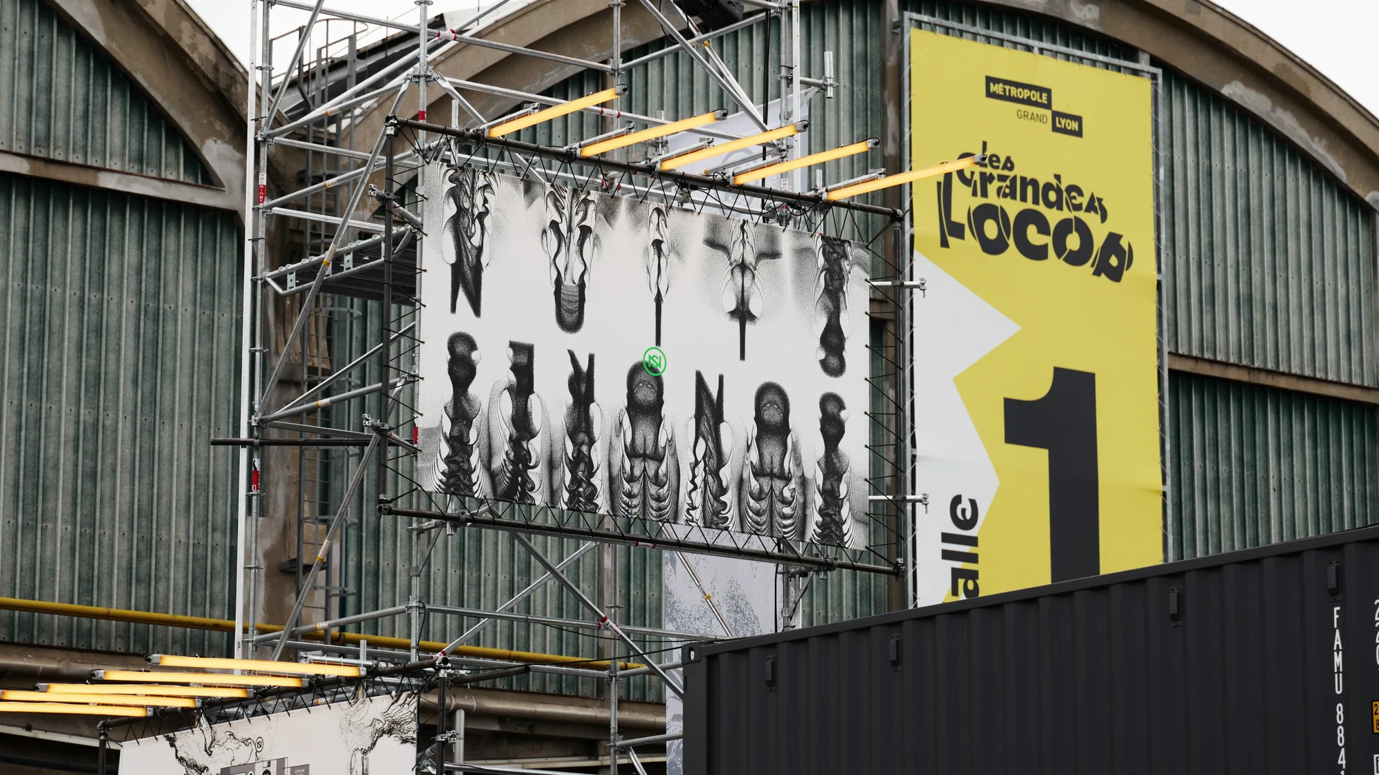 Billboard featuring black and white graphic art mounted on scaffolding, next to a large yellow sign with black text and graphics.