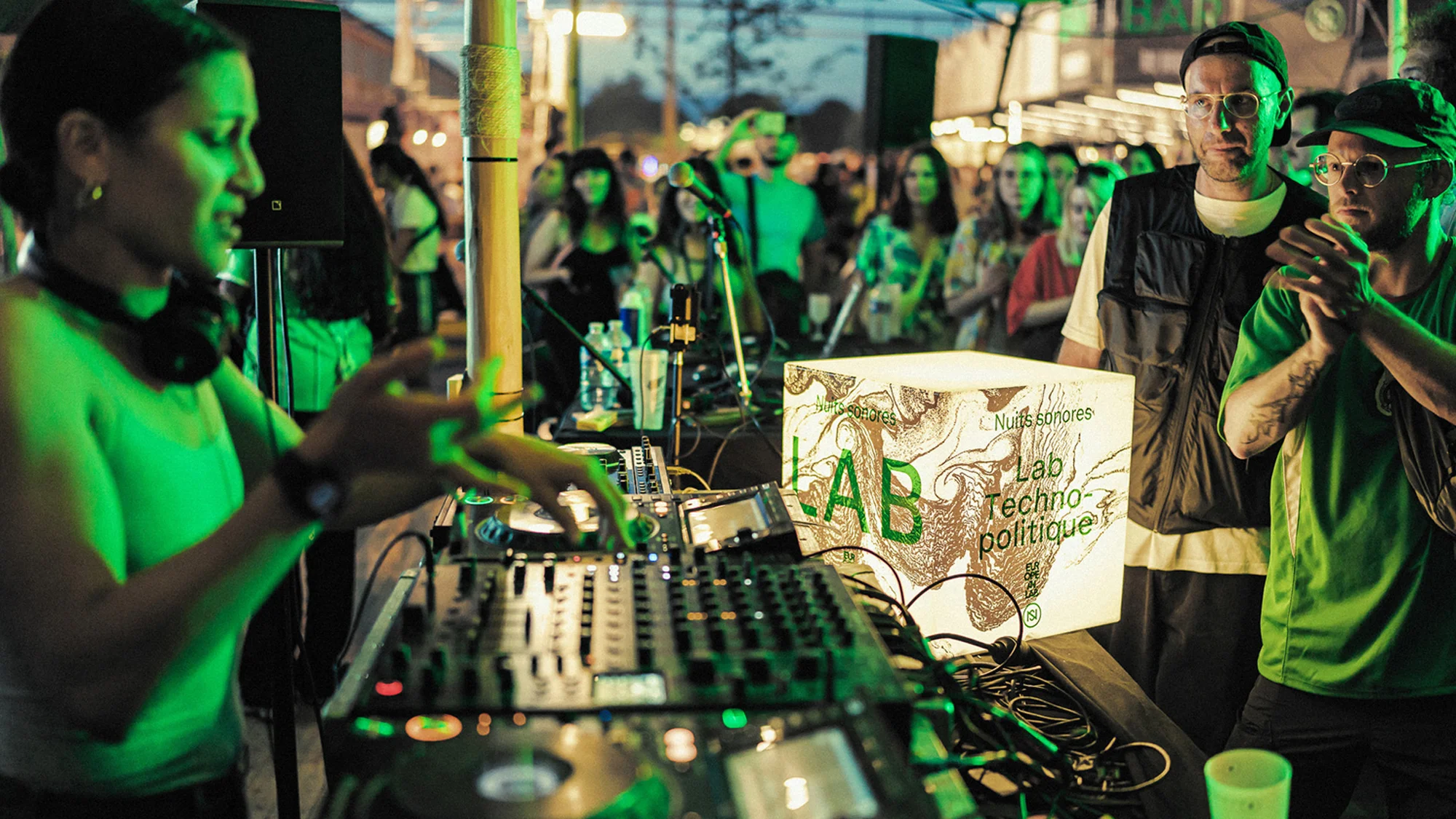 Female DJ mixing music at an outdoor event, with two men looking on and a crowd of people gathered in the background.