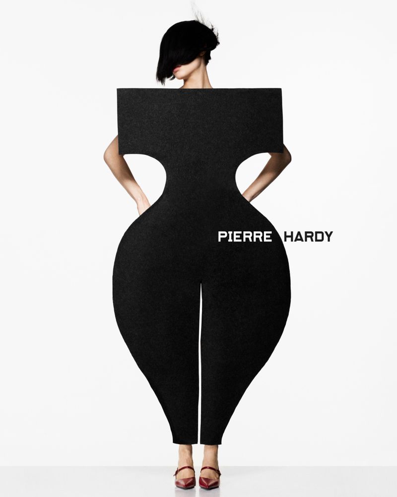 Fashion image features woman posing behind a large, black silhouette of an hourglass figure, styled with a dark bob haircut and red shoes.