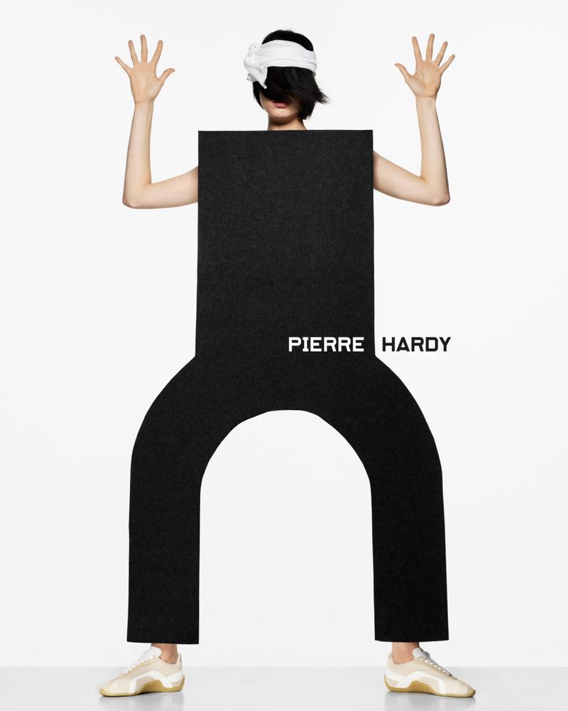 Fashion shoot featuring a person wearing a black rectangular prop, white headband, and beige sneakers in a minimalist studio.