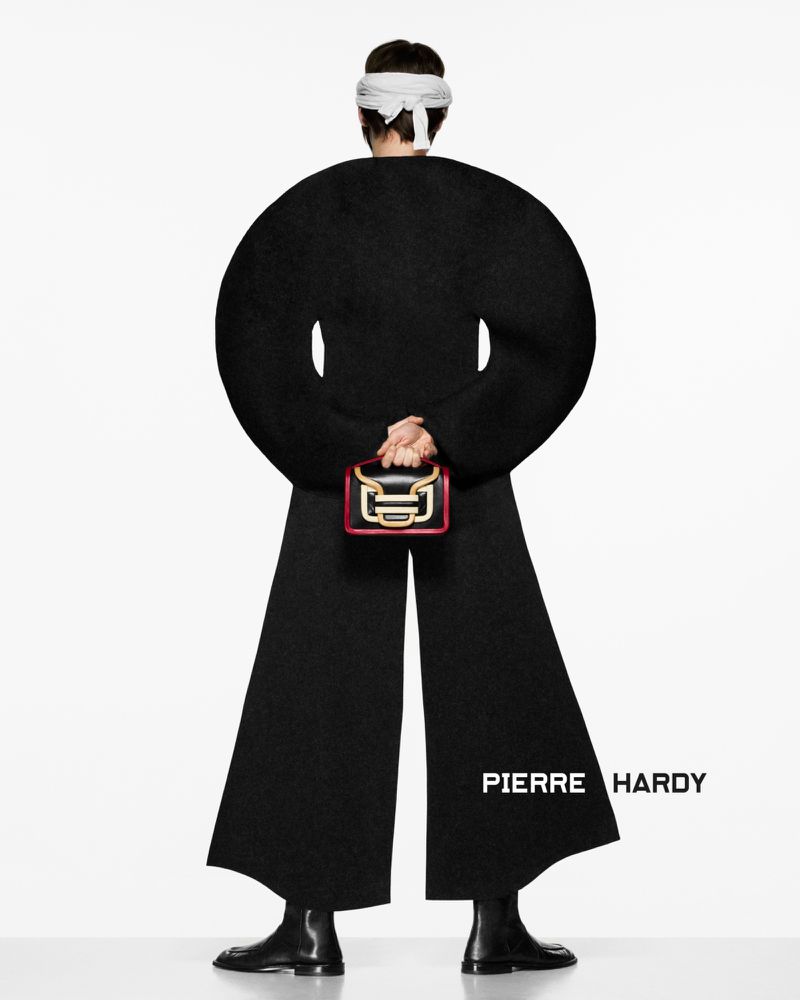 Back view of a model wearing a black outfit with a round silhouette, black boots, and a white head covering, holding a small purse.