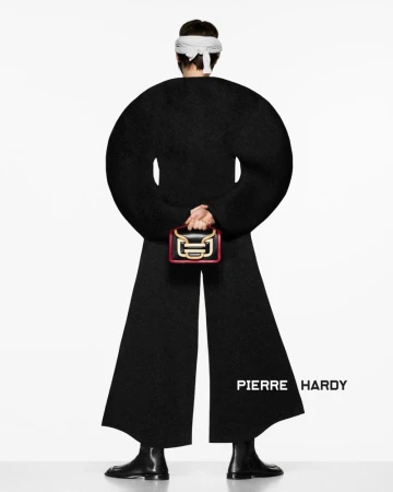 Back view of a model wearing a black outfit with a round silhouette, black boots, and a white head covering, holding a small purse.