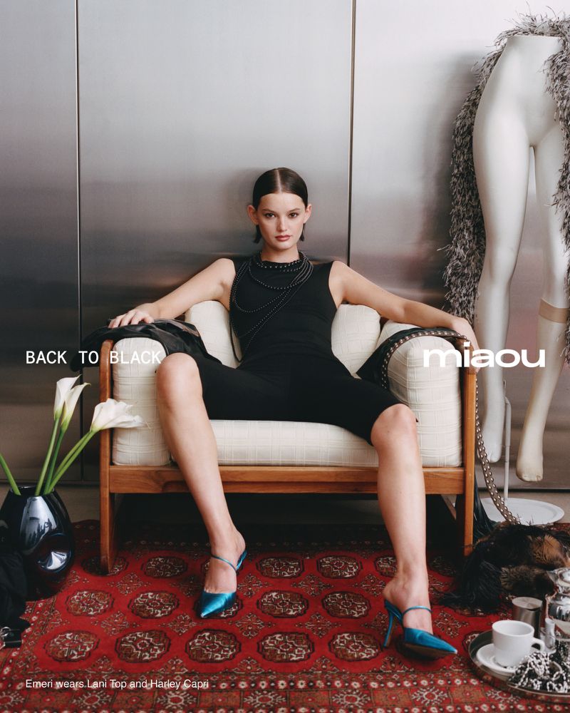 A slender woman with sleek hair sits in a cream chair in an indoor studio, wearing a black romper and teal mules for a fashion-forward look.