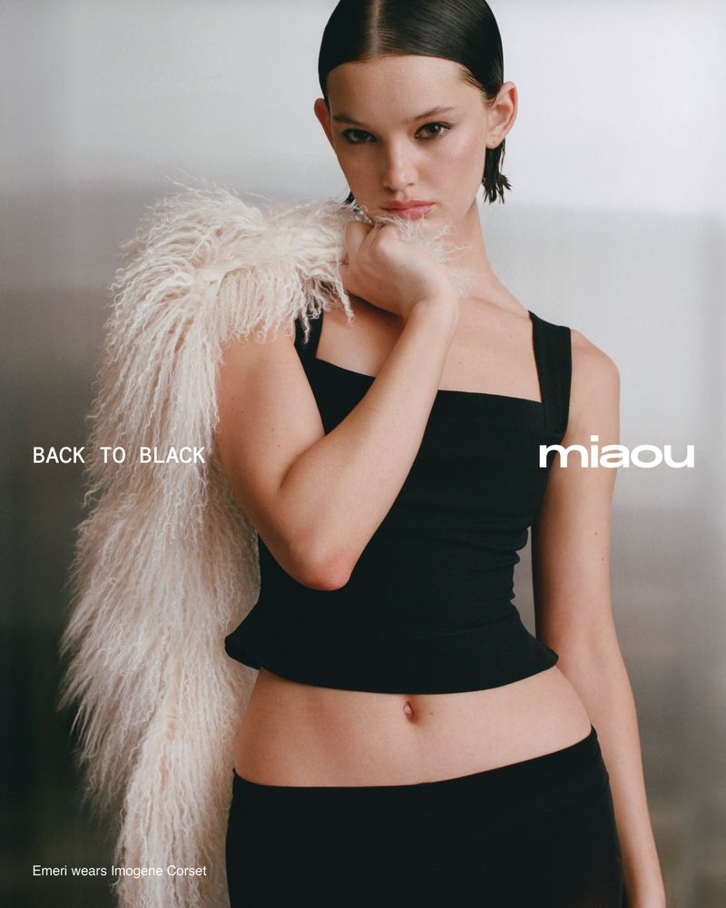 A young woman poses in a black top and skirt, with a furry white shawl draped over her shoulder, against a neutral backdrop.