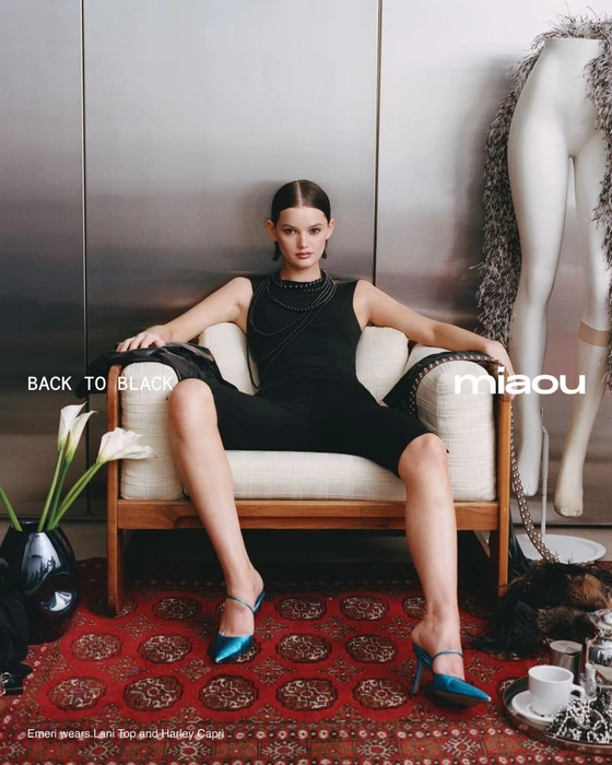 Model wearing black sleeveless top and blue heels seated in a cream armchair on a patterned red rug.
